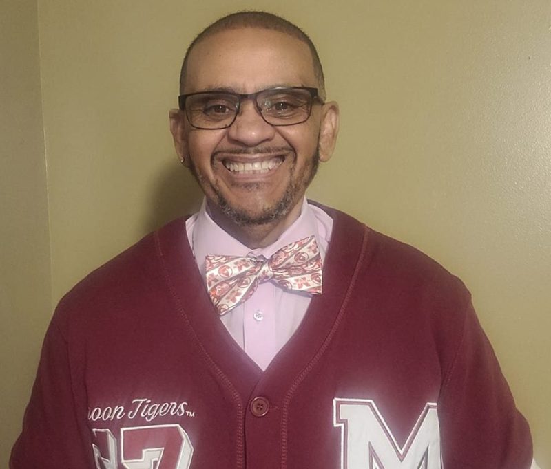 Charley Burton wears a maroon cardigan with white and red emblems over a pink button-down shirt and bowtie. He also wears black-rimmed glasses and smiles in front of a taupe background.